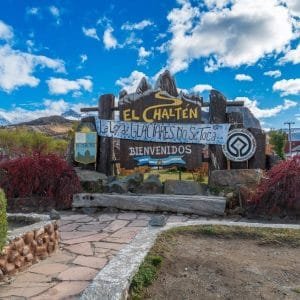 Cartel de madera de bienvenida a El Chaltén decorado con una bandera argentina bajo un cielo azul con nubes.