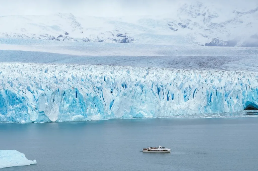 Barco de excursión navegando en las aguas del Lago Argentino frente a la imponente pared de hielo azul del Glaciar Perito Moreno.