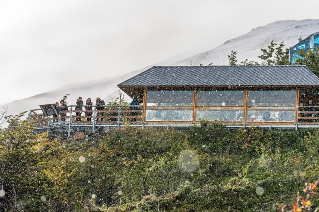 Turistas resguardados en un mirador de madera techado observando el paisaje del Parque Nacional Los Glaciares bajo una suave nevada.